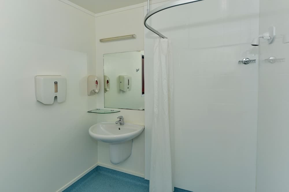 A clean, compact bathroom with white walls, a shower curtain, sink, mirror, and soap dispensers offers a functional space ideal for travelers seeking basic amenities in accommodations.