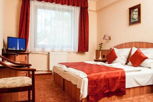A cozy hotel room features a neatly made bed with red accents, a small desk with a TV displaying a cityscape, warm lighting, and a window with red curtains, ideal for travelers seeking comfort and relaxation.