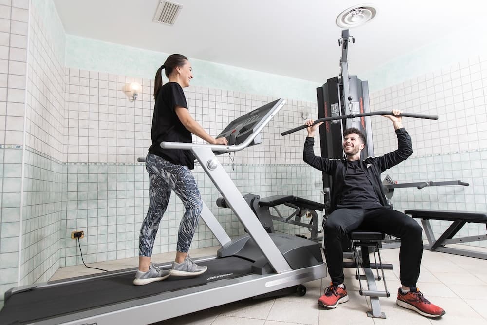 In a bright indoor gym with tiled walls, a woman on a treadmill and a man using a weight machine share a cheerful moment, showcasing fitness equipment ideal for travelers seeking active accommodations.
