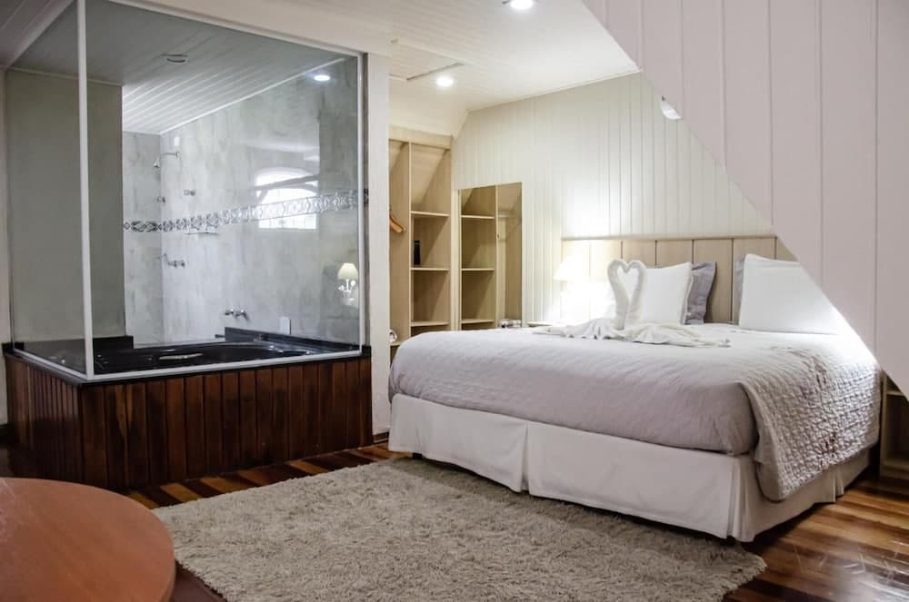 A cozy bedroom with a neatly made bed adorned with heart-shaped towels, a plush rug, wooden flooring, and an adjacent glass-enclosed bathroom featuring a luxurious bathtub, ideal for romantic getaways or spa retreats.