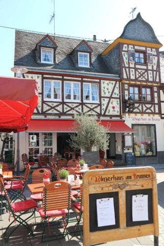 A charming European town square features a cozy outdoor café with red umbrellas and wooden chairs, set against a picturesque half-timbered building adorned with floral decorations, ideal for travelers seeking a quaint dining experience.