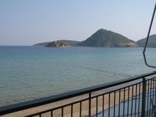 A serene coastal view from a balcony showcases tranquil turquoise waters, distant green hills, and a sandy beach, ideal for relaxing seaside getaways.