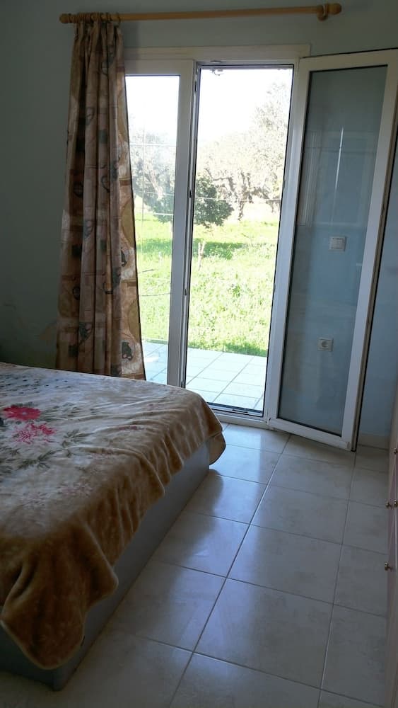 A cozy bedroom with tiled floors, floral bedding, and patterned curtains opens to a serene outdoor view through glass doors, ideal for travelers seeking a peaceful countryside retreat.