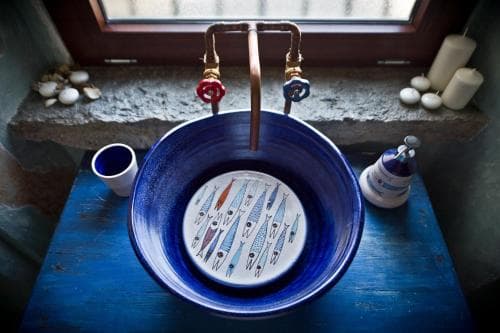 A charming rustic bathroom features a vibrant blue ceramic sink with fish designs, set against a stone windowsill adorned with candles and seashells, evoking a serene coastal retreat ideal for travelers seeking unique accommodations.