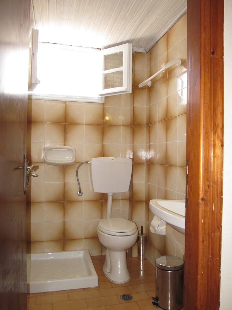 A cozy, compact bathroom with beige tiled walls, a small sink, toilet, bidet, and window, exudes a warm and functional atmosphere ideal for travelers seeking basic yet comfortable accommodations.
