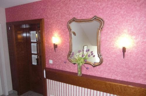 A cozy hotel hallway features pink textured walls, a vintage mirror, floral decorations, and warm sconce lighting, creating an inviting atmosphere ideal for travelers seeking a charming and comfortable stay.