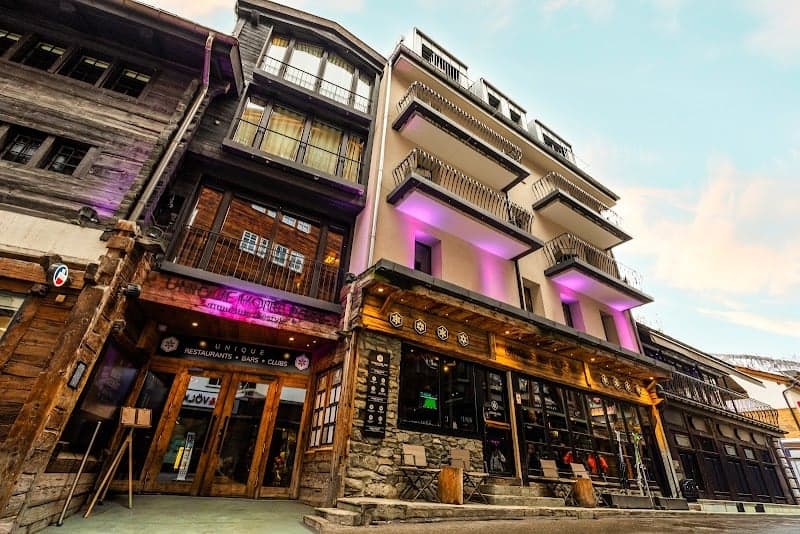 A vibrant evening scene at a charming hotel on Bahnhofstrasse in Zermatt, Switzerland, features modern architecture with purple lighting, inviting storefronts, and a cozy alpine atmosphere perfect for travelers seeking a luxurious mountain retreat.
