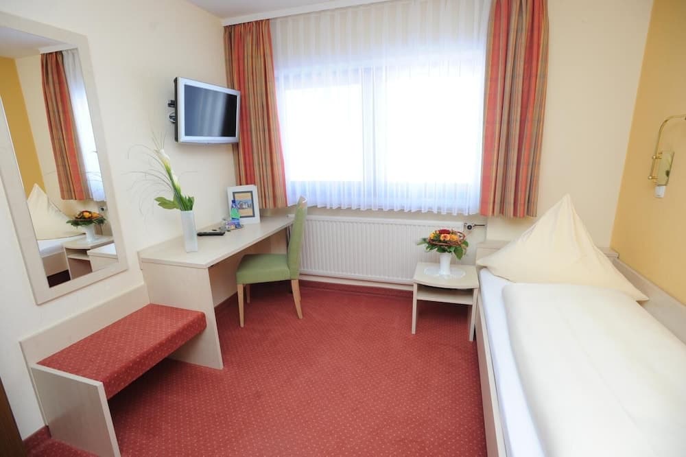 A cozy hotel room features a neatly made bed, a desk with a green chair, a wall-mounted TV, and a window with striped curtains, creating a welcoming atmosphere ideal for travelers seeking comfort and functionality.