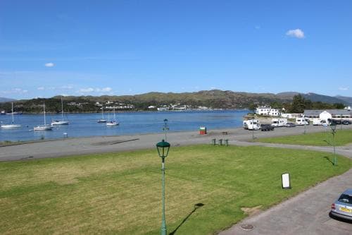 A serene coastal town scene features calm waters dotted with sailboats, lush green grassy areas, parked vehicles, and distant hills under a clear blue sky, ideal for a relaxing seaside getaway.
