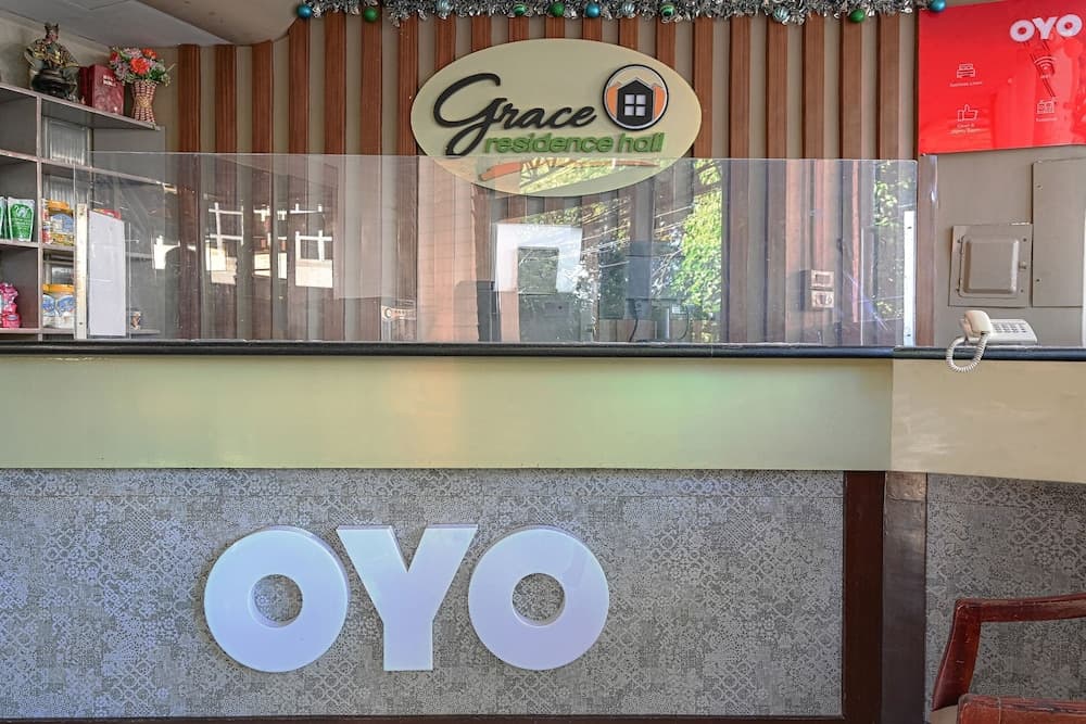 A cozy hotel reception at Grace residence hall features a protective glass barrier, vibrant decor, and an OYO logo, ideal for travelers seeking comfort and safety during their stay.