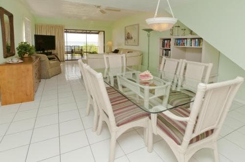 A cozy, well-lit beachside villa interior features a dining area with striped cushioned chairs around a glass table, a living room with a TV and bookshelf, and a sliding glass door leading to an outdoor balcony, ideal for a relaxing vacation retreat.