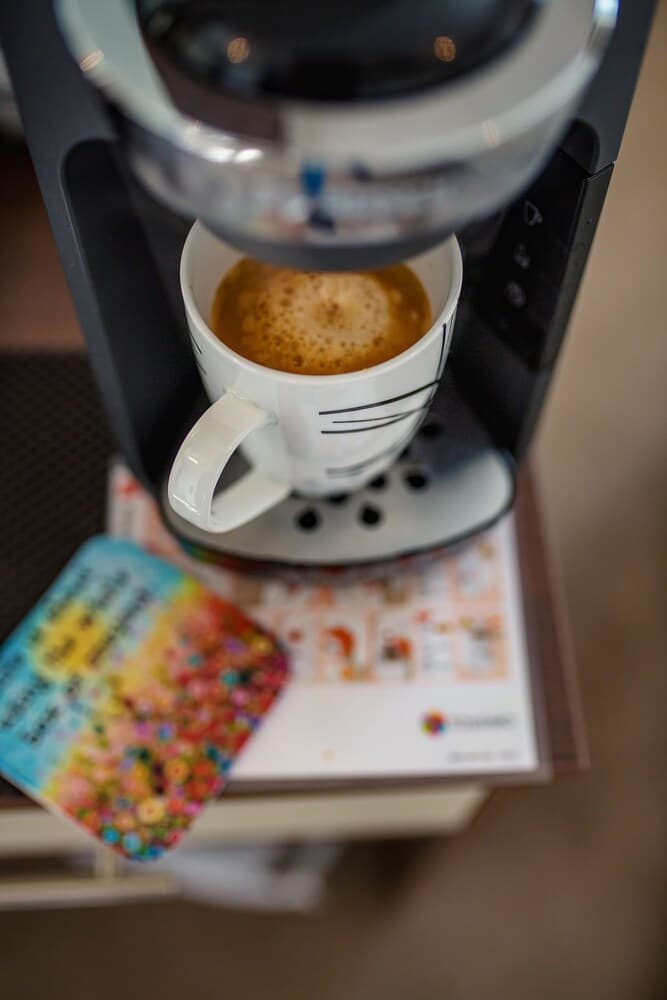 A cozy café scene features a freshly brewed coffee in a white mug under a sleek coffee machine, accompanied by colorful coasters on a wooden table, ideal for travelers seeking a relaxing coffee break.