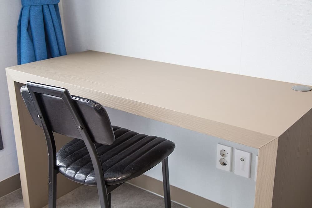 A minimalist hotel room workspace features a sleek wooden desk with built-in power outlets, a black cushioned chair, and a blue curtain backdrop, ideal for travelers seeking a functional and serene environment for work or study.