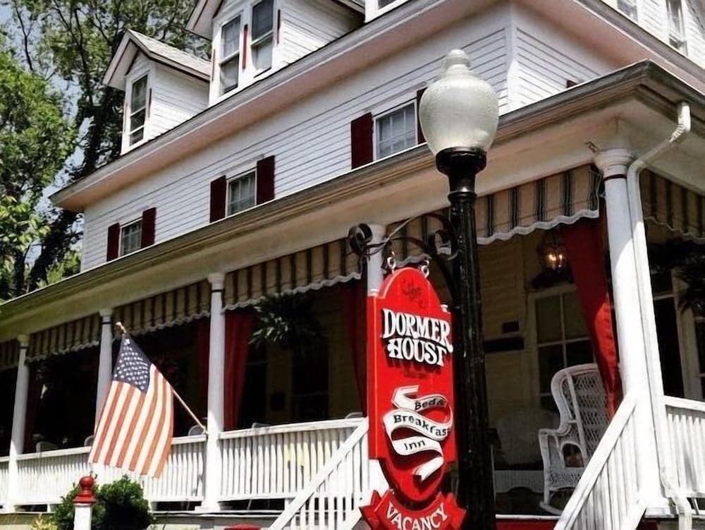 A charming white Victorian-style bed and breakfast with a red "Dormer House" sign, American flag, and inviting porch offers a cozy retreat for travelers seeking a quaint stay.