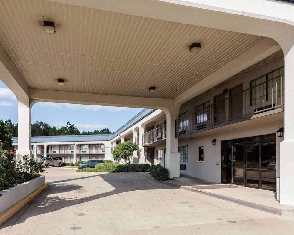 A serene motel courtyard features a covered driveway leading to a two-story building with balconies, lush greenery, and parked cars, ideal for travelers seeking a quiet and convenient stay.