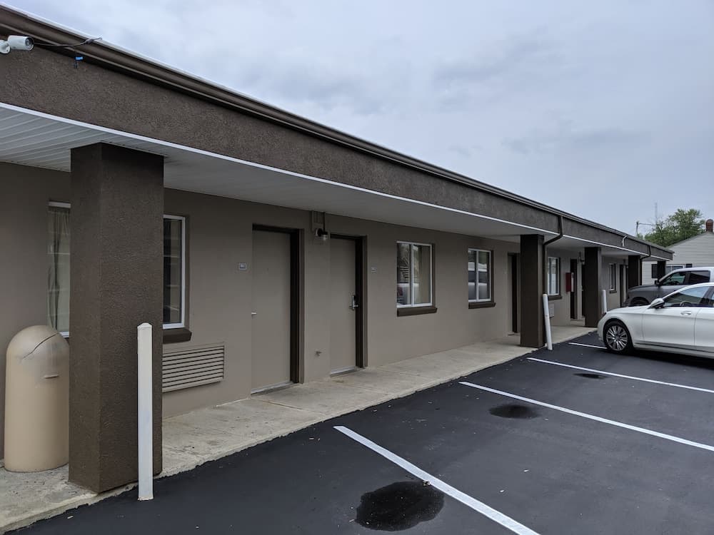 A quiet motel scene under an overcast sky features a row of beige rooms with black-framed windows and doors, a paved parking lot with marked spaces, and a parked white car, ideal for travelers seeking budget-friendly accommodations.
