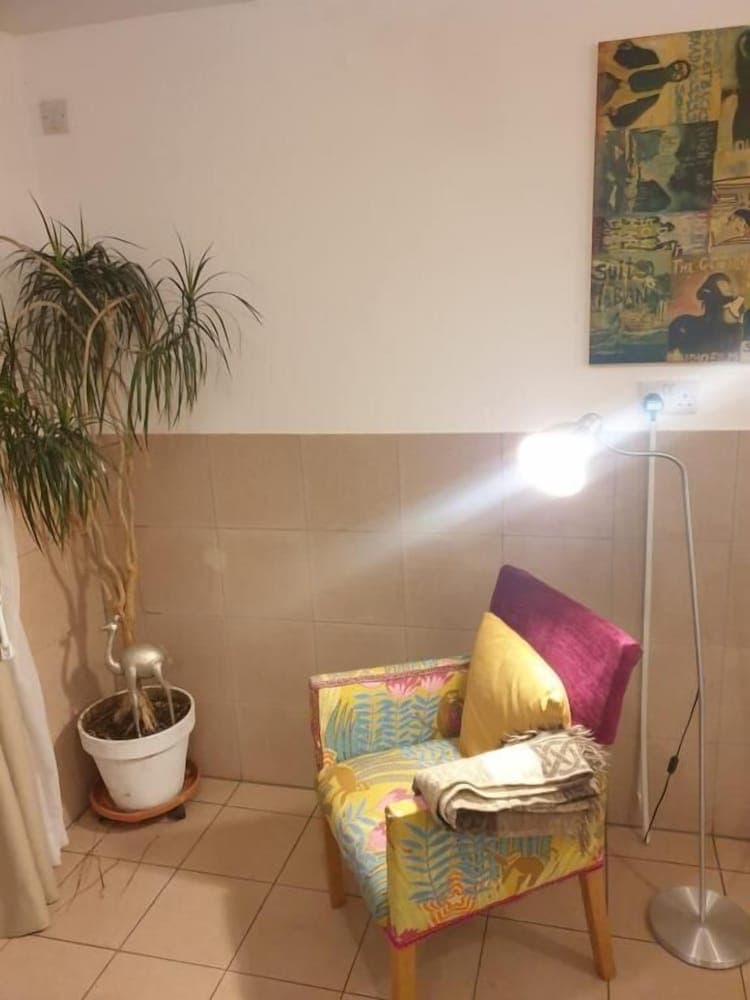 A cozy corner in a tiled room features a vibrant armchair adorned with colorful cushions, a bright floor lamp, a potted plant, and wall art, creating an inviting atmosphere perfect for travelers seeking a comfortable reading nook.