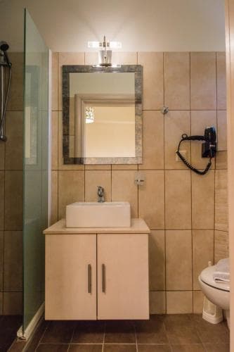 A cozy hotel bathroom features beige tiled walls, a sleek vanity with a white sink, a framed mirror, and a glass shower partition, ideal for travelers seeking comfort and functionality.