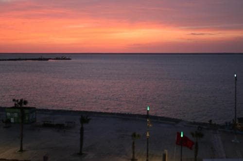 A serene beachfront at sunset showcases vibrant orange and pink skies over calm waters, with palm trees, a distant ship, and a Turkish flag enhancing the tranquil seaside atmosphere ideal for relaxation and photography.