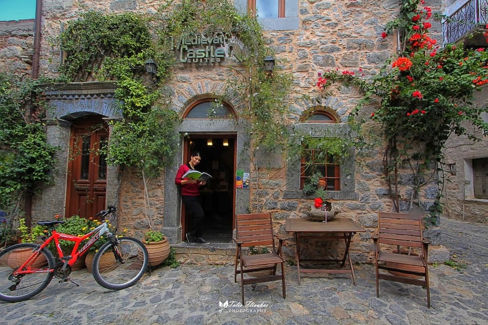 A cozy medieval stone building adorned with ivy and vibrant flowers invites travelers to relax at its rustic outdoor seating area, perfect for a serene getaway or a cyclist's rest stop.