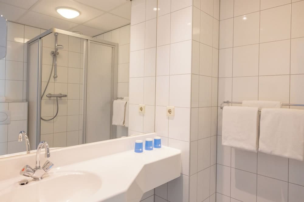 A clean, modern hotel bathroom features white tiled walls, a sleek sink with dual faucets, a shower enclosure, neatly folded towels, and small blue toiletries, ideal for travelers seeking comfort and hygiene.