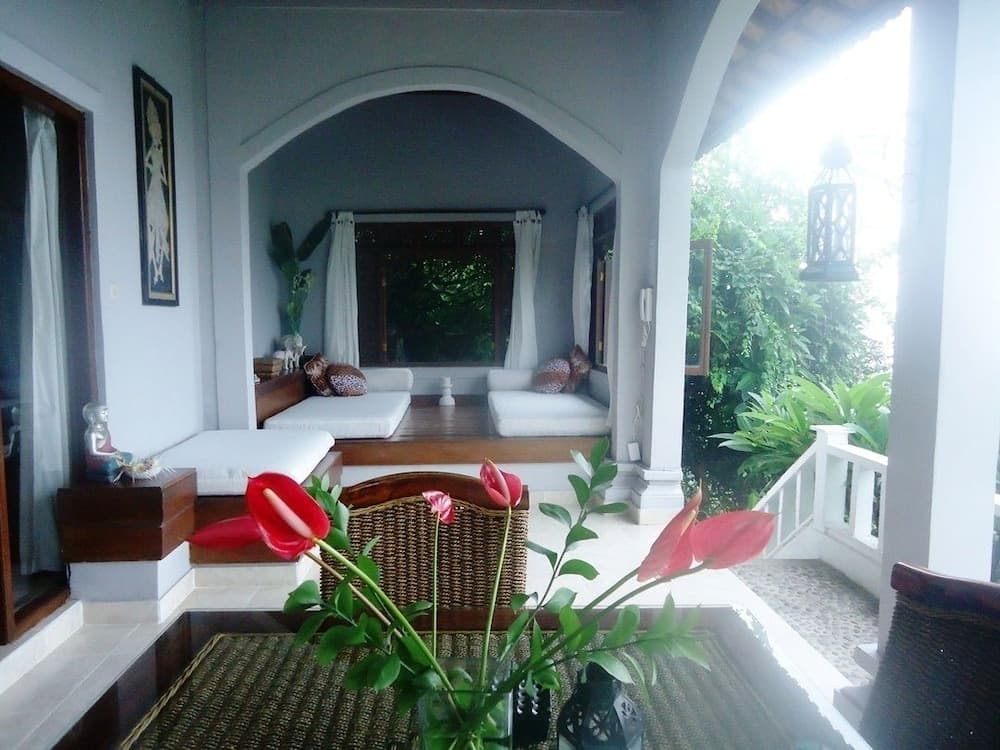 A serene tropical villa balcony features plush seating, vibrant red flowers, lush greenery outside, and a tranquil ambiance ideal for relaxation and leisure travel.