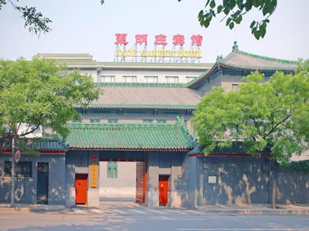 A serene entrance to the Jade Garden Hotel features traditional Chinese architecture with green-tiled roofs, red doors, and lush trees, ideal for travelers seeking cultural immersion and relaxation.