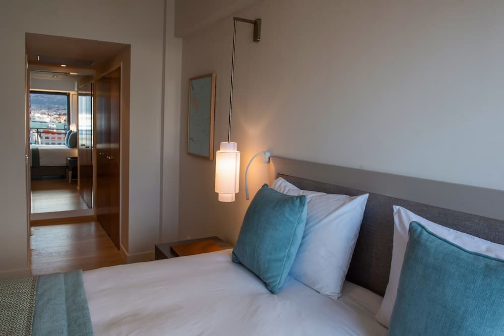 A cozy hotel bedroom with teal pillows, modern wall lamp, and an open door leading to a balcony with scenic views, ideal for travelers seeking comfort and relaxation.