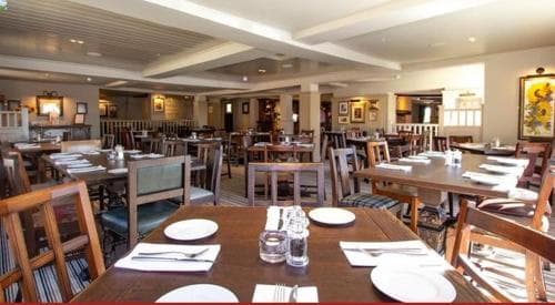 A cozy restaurant interior with neatly arranged wooden tables and chairs, warm lighting, and framed artwork creates an inviting atmosphere perfect for travelers seeking a comfortable dining experience.