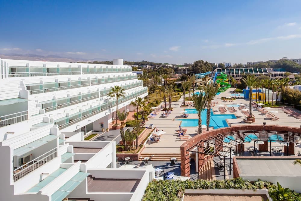 A vibrant resort scene featuring a sprawling white hotel complex, lush palm trees, colorful water slides, and a serene pool area under a clear blue sky, ideal for family vacations seeking relaxation and entertainment.