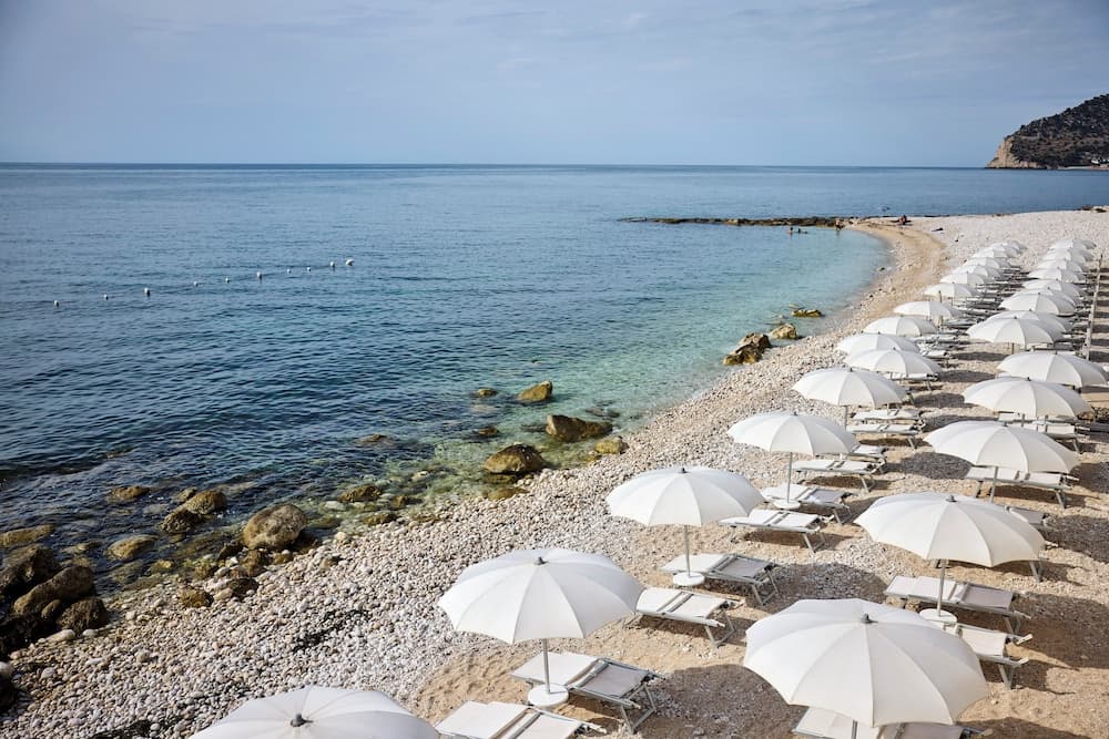A serene pebble beach lined with white umbrellas and lounge chairs invites relaxation under clear skies, perfect for a tranquil seaside getaway.