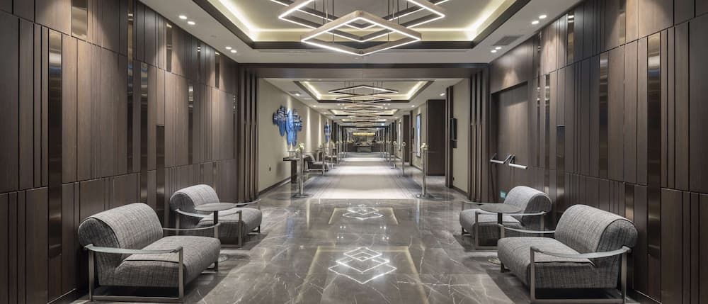 A luxurious hotel corridor features sleek wooden walls, modern geometric lighting, plush seating areas, and a polished marble floor, creating an inviting atmosphere ideal for travelers seeking comfort and elegance.