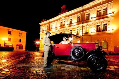 A vintage red car glows under warm streetlights on a cobblestone square at night, evoking nostalgia in front of illuminated historic buildings, perfect for a romantic getaway or history-themed travel photography.