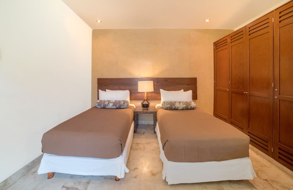 A cozy hotel room features two neatly made twin beds with brown linens, a wooden headboard, a central lamp on a small table, and a large wooden wardrobe, ideal for travelers seeking comfort and simplicity.
