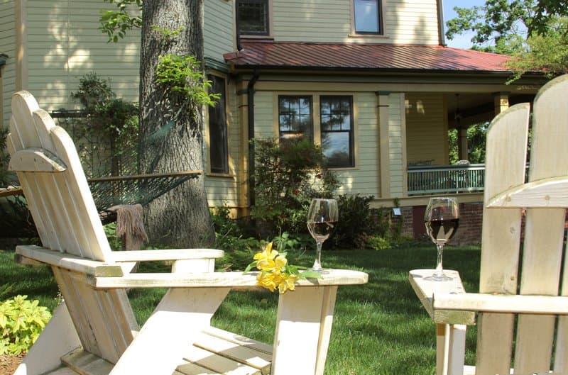 A serene suburban backyard features Adirondack chairs with wine glasses and yellow flowers on a sunny day, ideal for relaxing getaways or leisurely weekend retreats.
