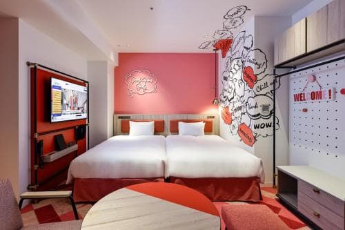 A vibrant hotel room features twin beds against a pink wall adorned with playful comic-style artwork, creating a cheerful and modern atmosphere ideal for travelers seeking a fun and cozy stay.