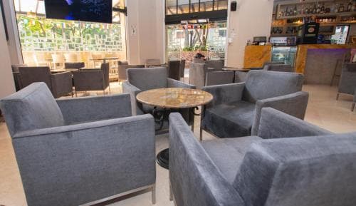 A cozy lounge area with plush gray armchairs and a round wooden table invites relaxation, set against a backdrop of vibrant greenery and a well-stocked bar, ideal for travelers seeking a serene spot to unwind during layovers or breaks.