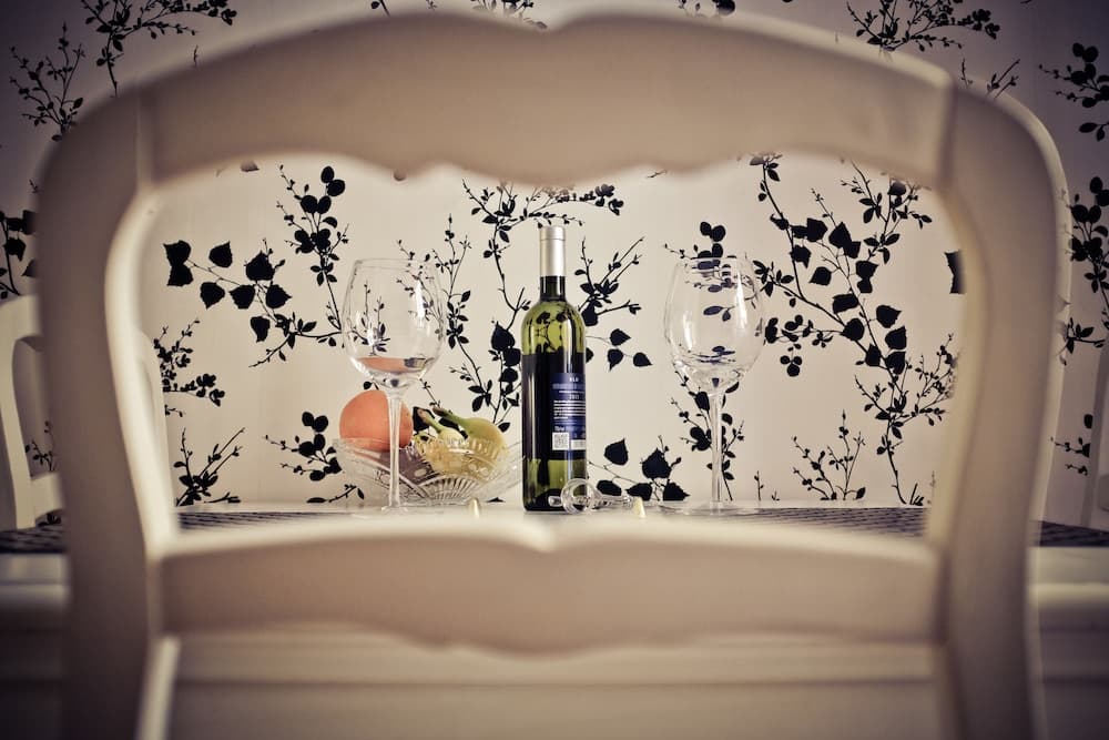 A cozy dining setup framed by a chair backrest features a bottle of wine, two glasses, and fruit on a table against a floral wallpaper backdrop, ideal for intimate dinner scenes or travel blog content showcasing charming indoor settings.