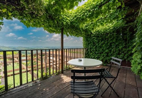 A serene balcony draped in lush greenery offers a picturesque view of rolling hills, perfect for a tranquil getaway with its cozy table and chairs set against a vibrant outdoor backdrop.