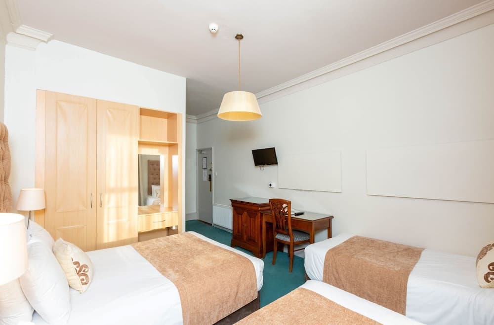 A cozy hotel room features two neatly made beds with beige throws, a wooden desk with a chair, a built-in wardrobe with a mirror, and soft lighting, ideal for travelers seeking comfort and functionality.