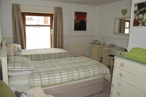 A cozy bedroom with twin beds adorned in green plaid bedding, featuring soft natural light from a window, a vintage-style dresser, and framed artwork, ideal for a serene getaway or family accommodation.