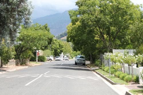 A serene suburban street lined with lush green trees and white fences leads towards distant mountains under a partly cloudy sky, ideal for a peaceful road trip or exploring nature's beauty.
