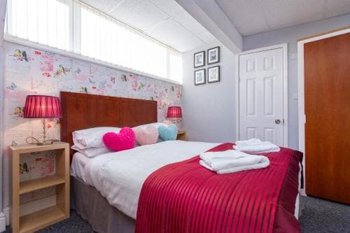 A cozy bedroom with a neatly made bed adorned in red and white linens, framed by patterned wallpaper and soft lighting from bedside lamps, creates an inviting atmosphere ideal for travelers seeking a comfortable stay.
