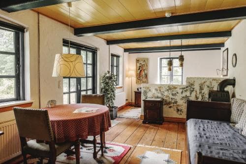 A cozy, rustic living room with wooden beams, a round table draped in a red cloth, and large windows inviting natural light, ideal for travelers seeking a charming countryside retreat.