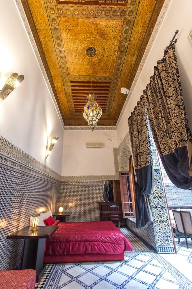 A cozy Moroccan-style bedroom with intricate tiled walls, ornate ceiling patterns, and vibrant red bedding invites travelers to experience luxurious relaxation in a traditional setting.