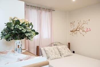A cozy bedroom with soft pink curtains, a floral wall decal, and a vibrant potted plant creates a serene atmosphere, ideal for travelers seeking relaxation and comfort.
