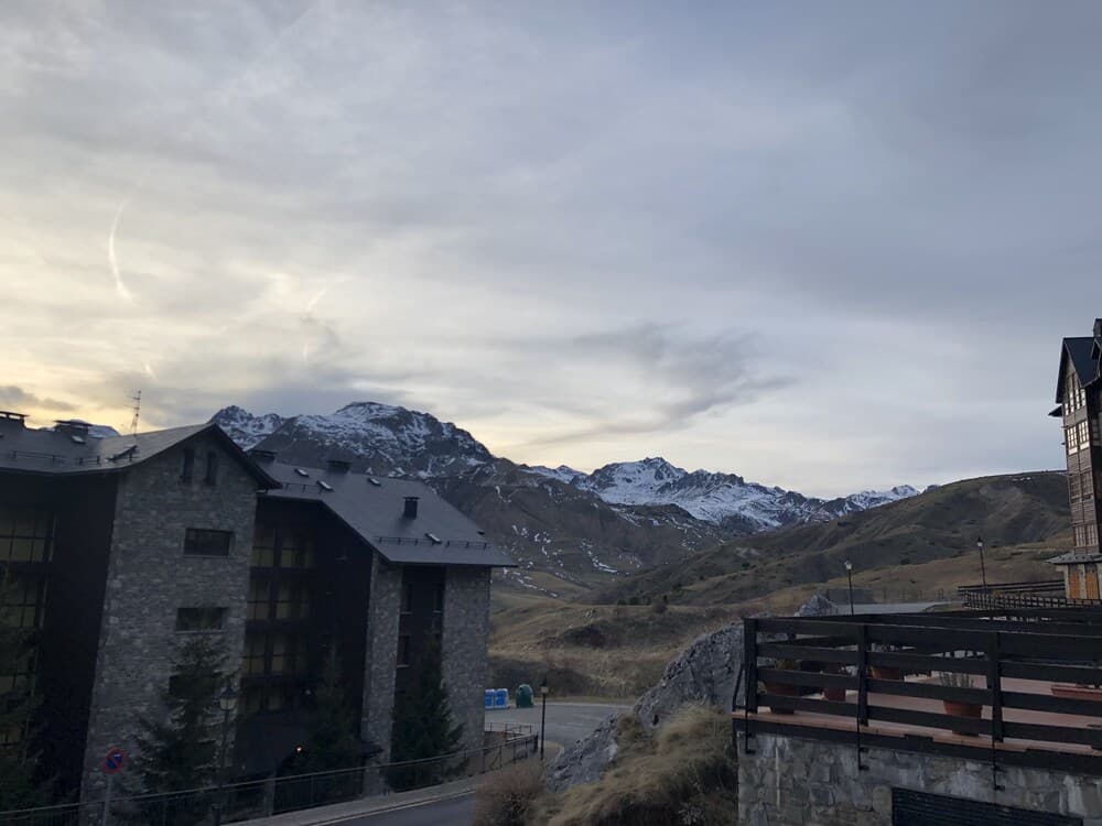 A serene mountain village scene features stone buildings under a cloudy sky, with snow-capped peaks in the background, ideal for travelers seeking peaceful alpine retreats.