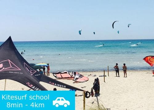 A vibrant kitesurf school on a sunny beach with colorful kites soaring above the turquoise sea, ideal for adventure seekers looking for an 8-minute drive or 4km trip to learn kitesurfing.