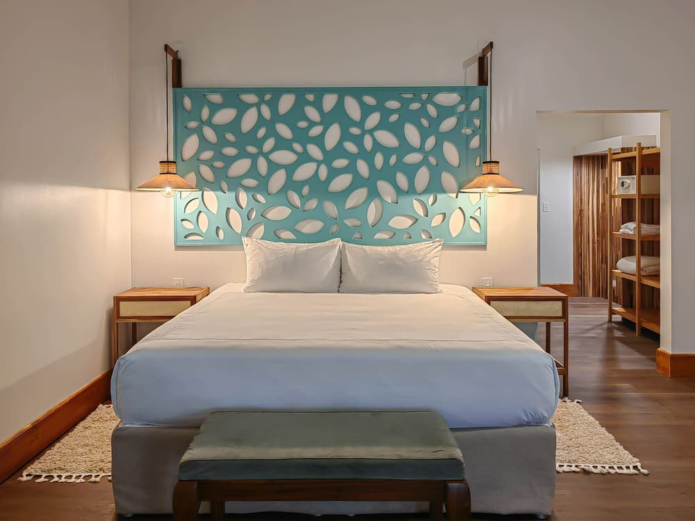 A serene hotel bedroom features a cozy bed with a unique teal leaf-patterned headboard, warm pendant lights, wooden furnishings, and a soft rug, ideal for travelers seeking relaxation and comfort.
