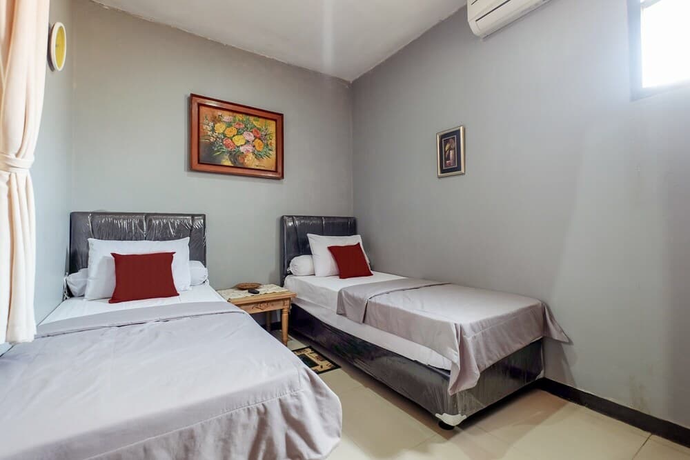 A cozy budget-friendly hotel room features two neatly made single beds with red accents, soft lighting from a window, and simple wall art, ideal for travelers seeking comfort and affordability.